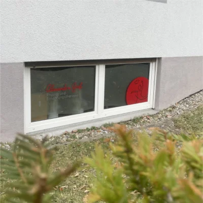 Ein Kellerfenster zeigt roten Text und ein rotes Logo und wirbt für die Leistungen von Physiotherapie-Termin. Im Vordergrund sind Gras und Felsen zu sehen. Das Fenster gehört zu einer Praxis für Physiotherapie und Ernährungstherapie.