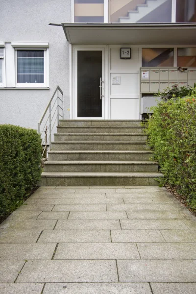 Eingang zu einem Mehrfamilienhaus mit Glastür, Stufen, Briefkästen auf der rechten Seite und Büschen am Gehweg – perfekt für Ihren nächsten Physiotherapie-Termin.