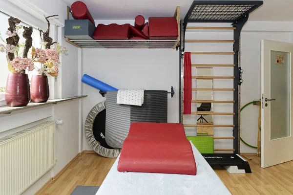 Ein Physiotherapieraum mit Gymnastikmatten, einem Trampolin, einem Sprossenreck, Schaumstoffrollen und rot gepolsterten Geräten für Physiotherapie-Behandlungen, mit Blumen in Vasen auf der Fensterbank.